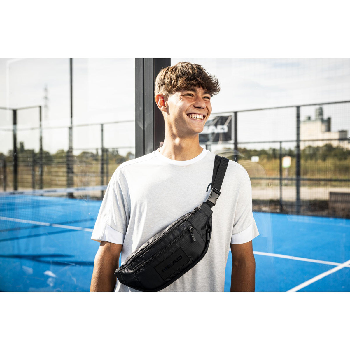 Smiling young man wearing a black HEAD crossbody waist pack over a white T-shirt on an outdoor tennis court, with adjustable strap and zippered compartments visible