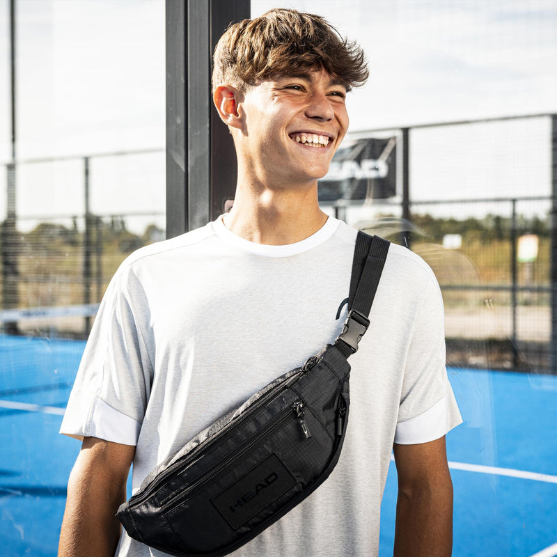 Smiling young man wearing a black HEAD crossbody waist pack over a white T-shirt on an outdoor tennis court, with adjustable strap and zippered compartments visible