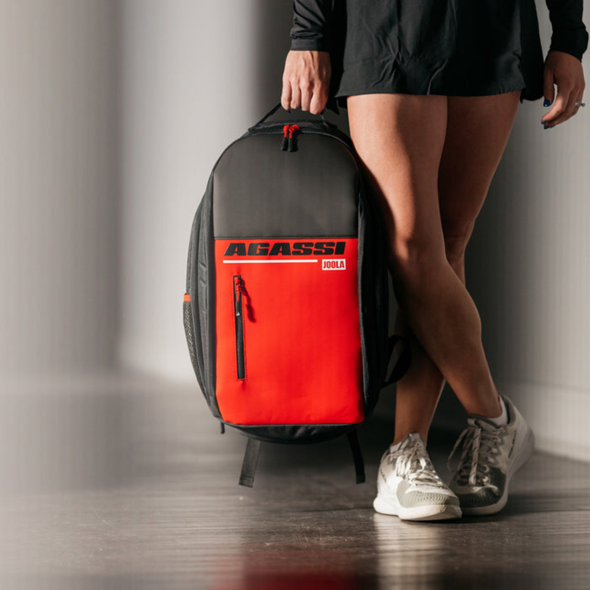Person holding a red and black Agassi tennis bag against a neutral background