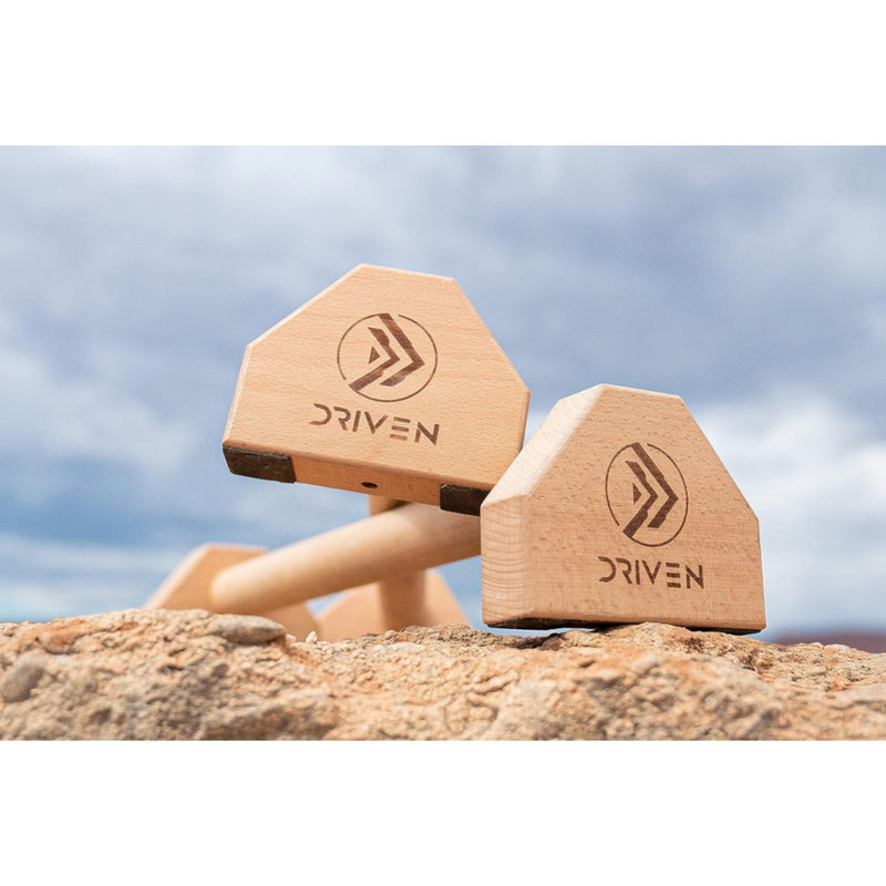 Wooden dumbbells with 'Driven' branding on a rocky surface against a cloudy sky.