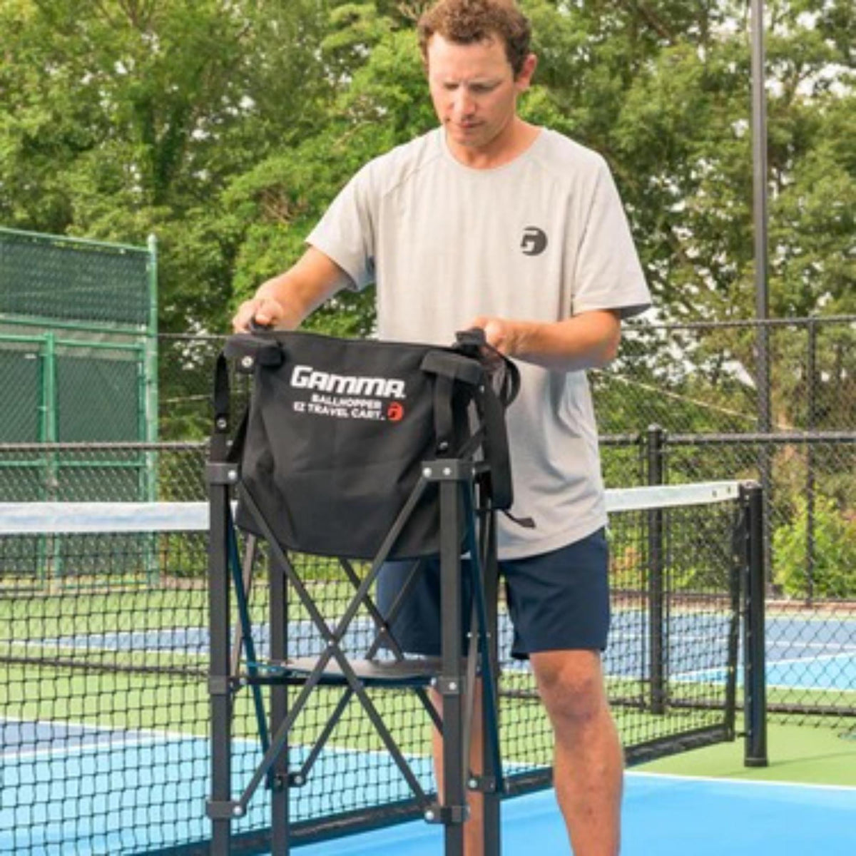 Portable ball hopper cart with rolling wheels, collapsible frame, and attached storage basket for transporting sports balls.