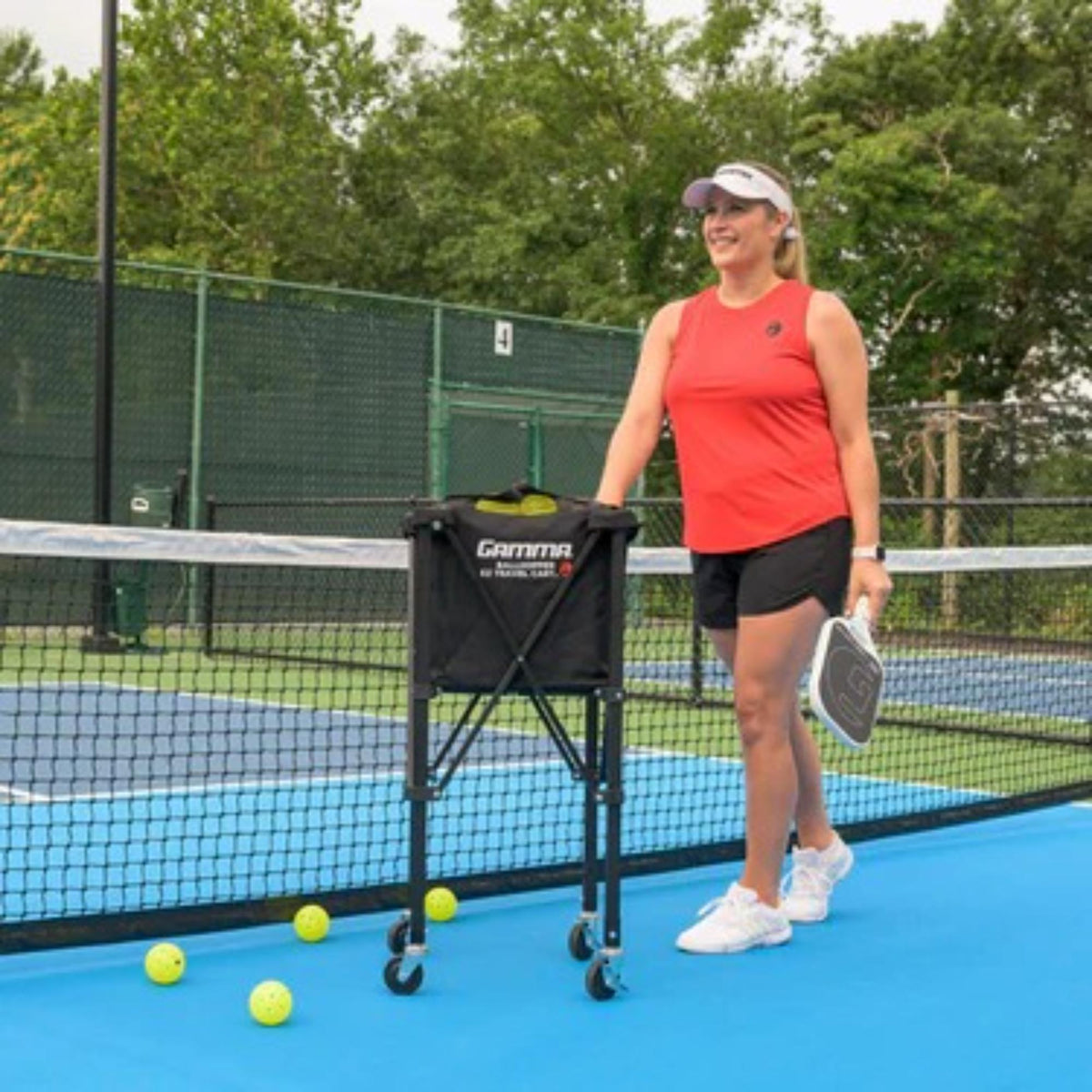 Portable ball hopper cart with rolling wheels, collapsible frame, and attached storage basket for transporting sports balls.