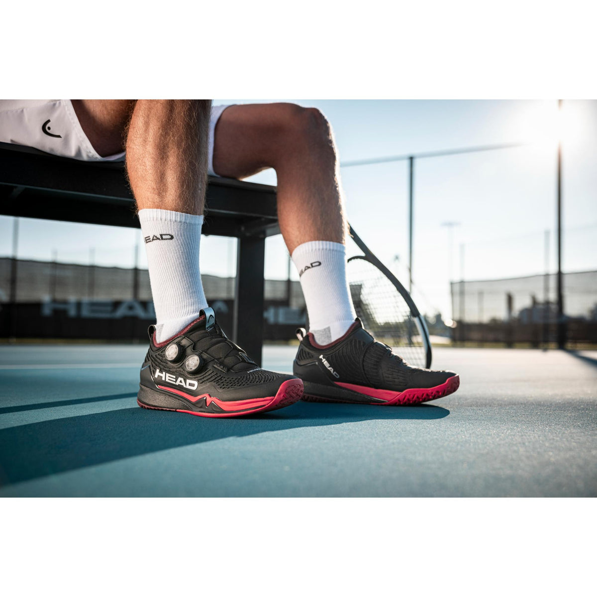 Man wearing black and red HEAD Endure Pro BOA tennis shoes while sitting on a court bench, shown in an outdoor tennis court setting with a racket nearby.
