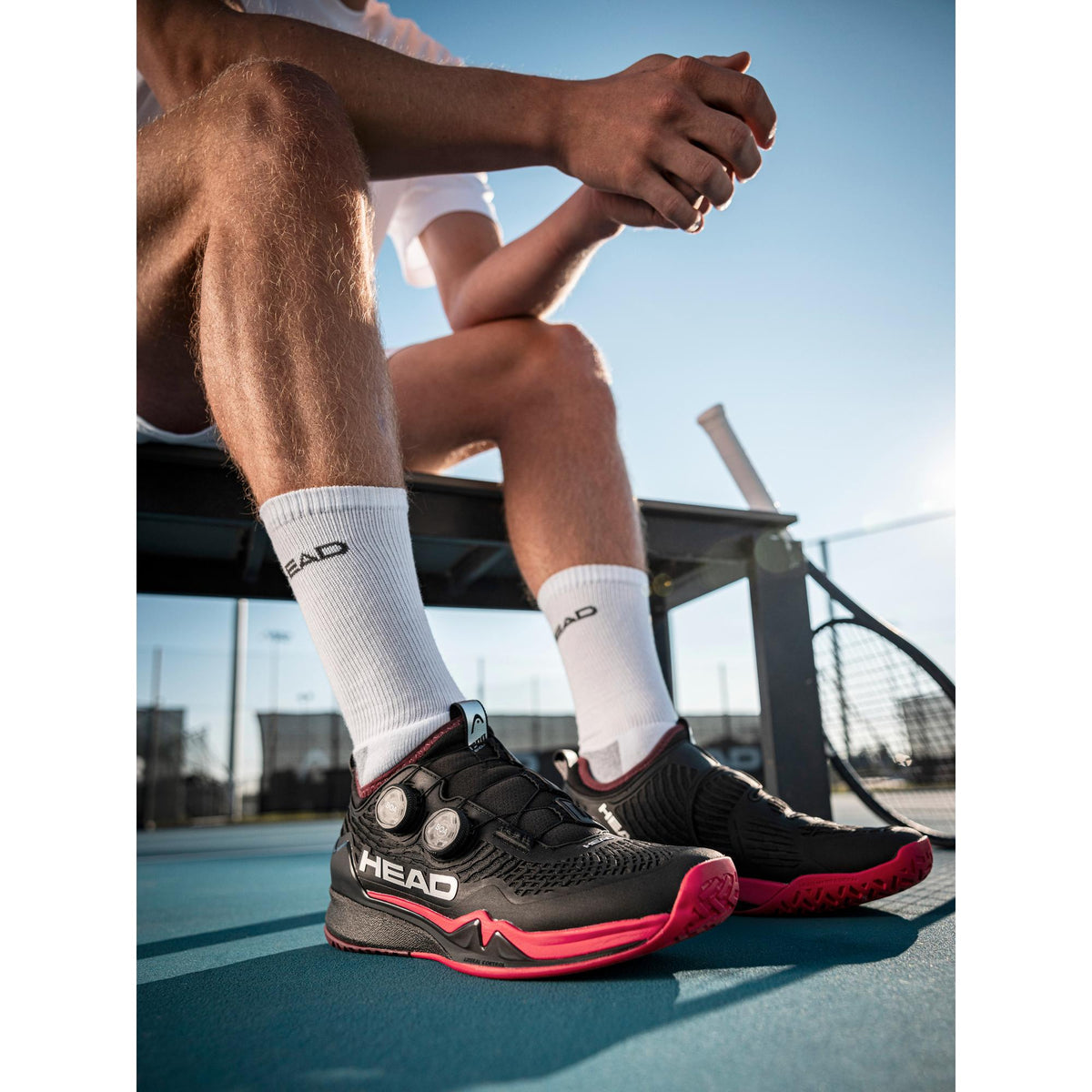 Man wearing black and red HEAD Endure Pro BOA tennis shoes while sitting on a court bench, shown in an outdoor tennis court setting with a racket nearby.