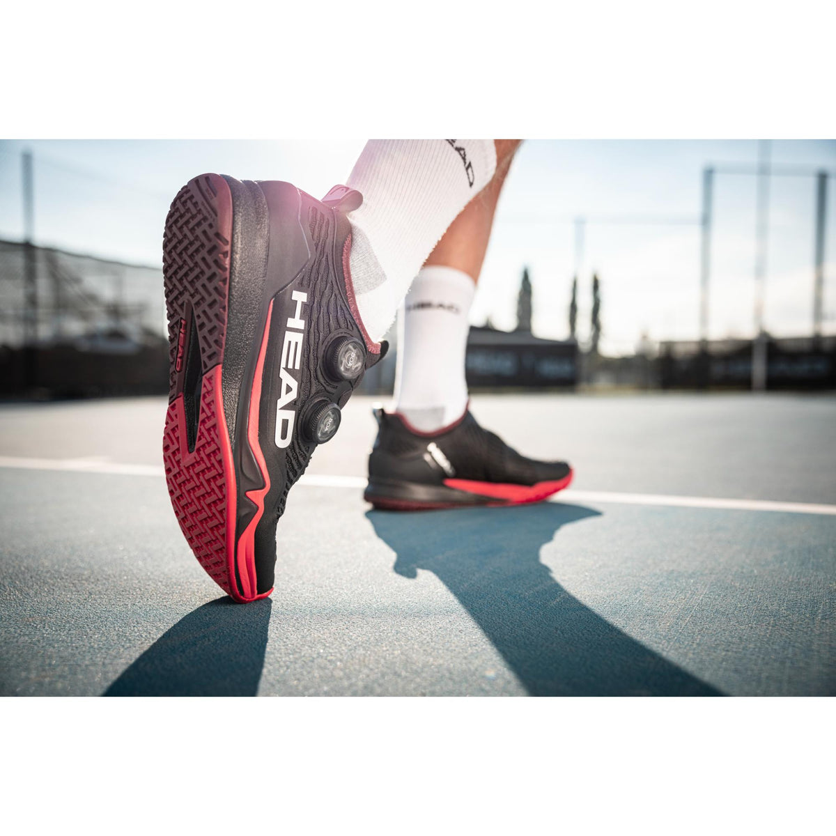 Close-up of a person wearing black and red HEAD Endure Pro BOA tennis shoes while stepping on an outdoor tennis court, highlighting the outsole grip and BOA dial system.