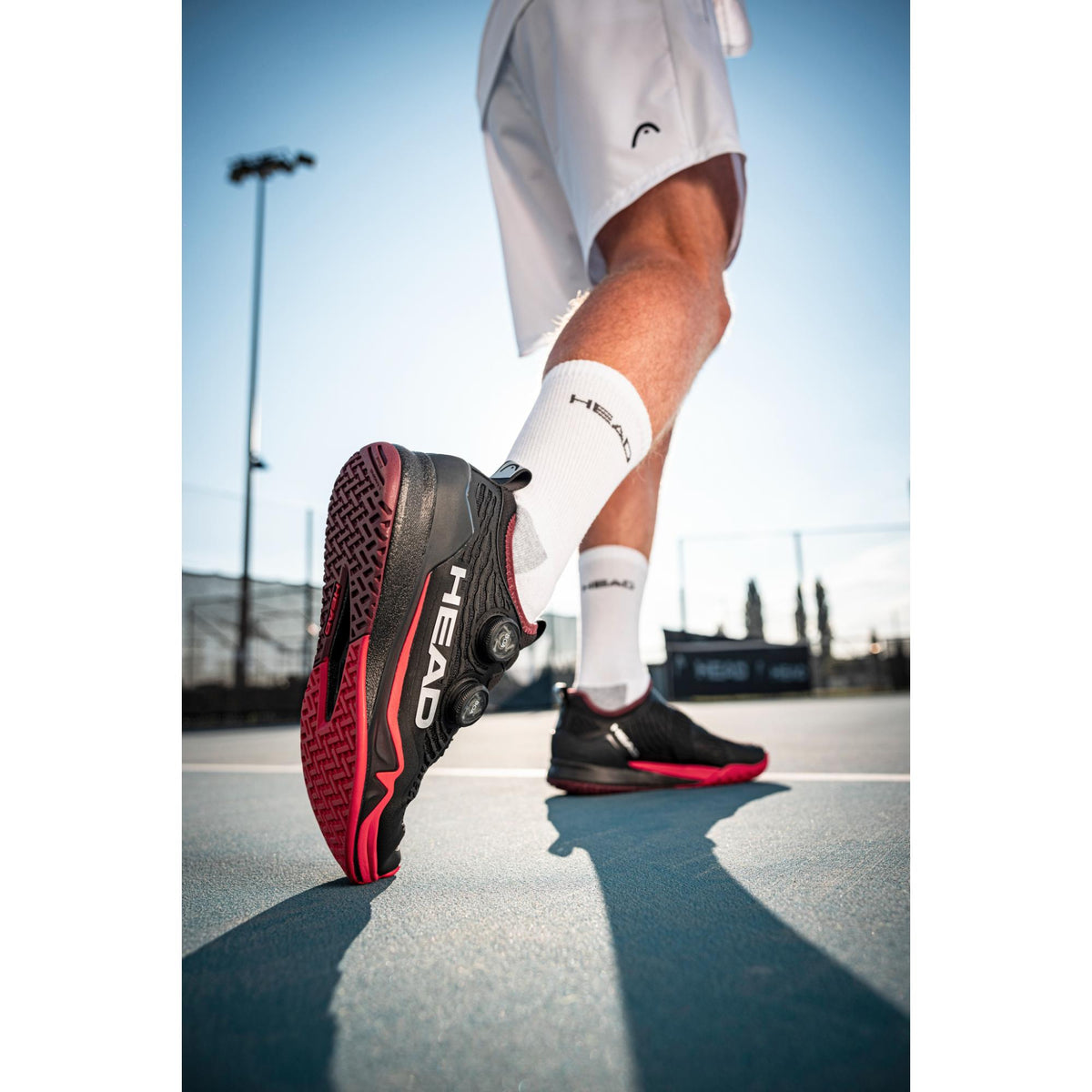 Close-up of a person wearing black and red HEAD Endure Pro BOA tennis shoes while stepping on an outdoor tennis court, highlighting the outsole grip and BOA dial system.