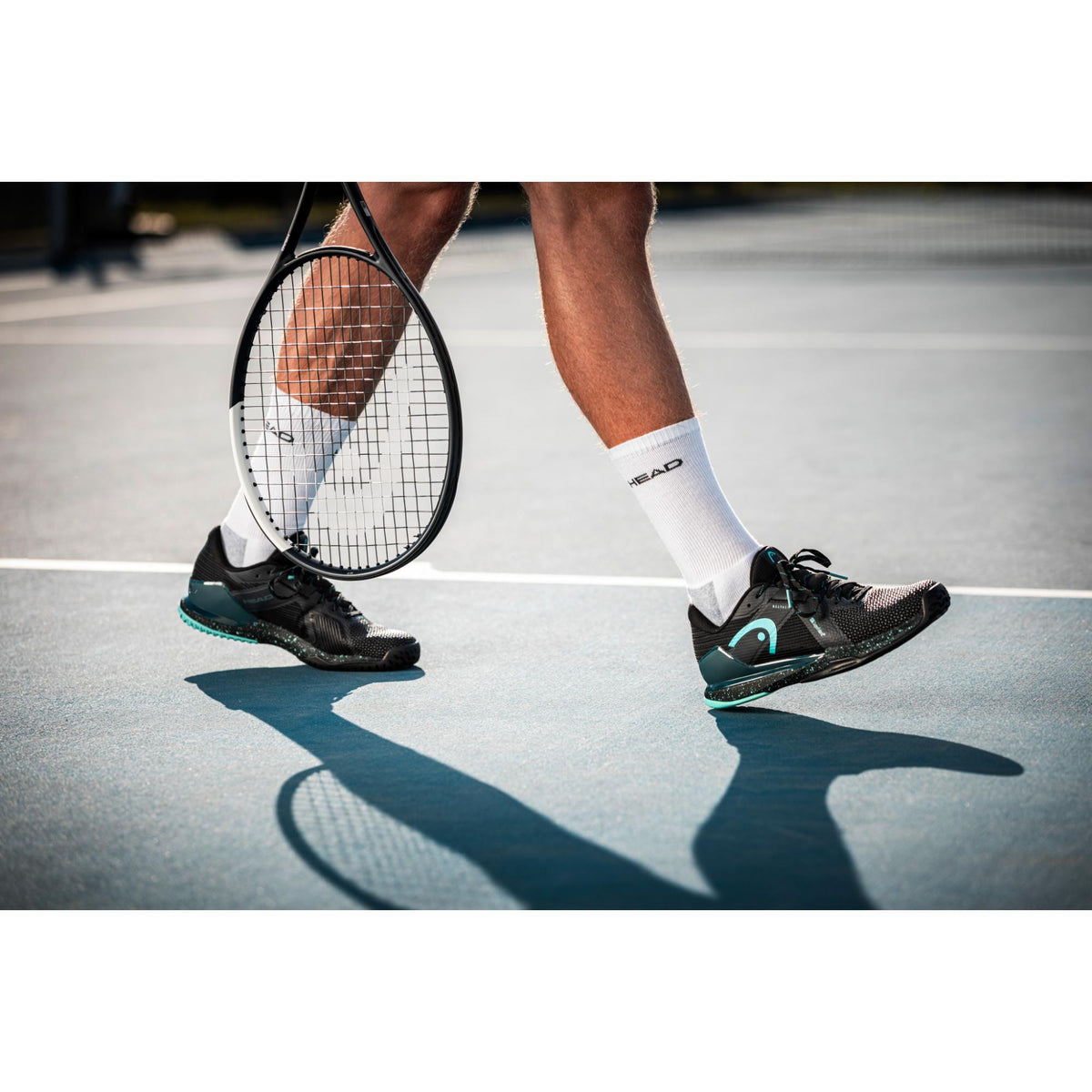 Tennis player walking on a hard court wearing black and teal HEAD Sprint Pro 4.0 SF tennis shoes, holding a racket at their side, with focus on footwork, shoe cushioning, and court surface.