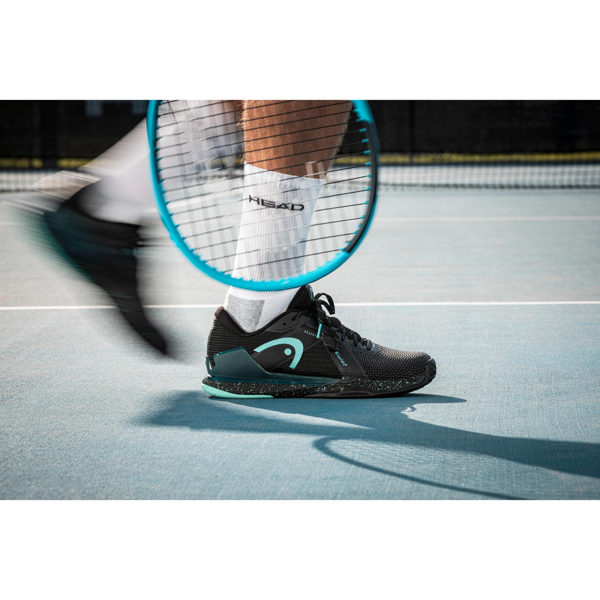 Tennis player in motion on a hard court wearing black and teal HEAD Sprint Pro 4.0 SF tennis shoes, with a racket swinging past the frame and focus on stability, traction, and on-court performance.