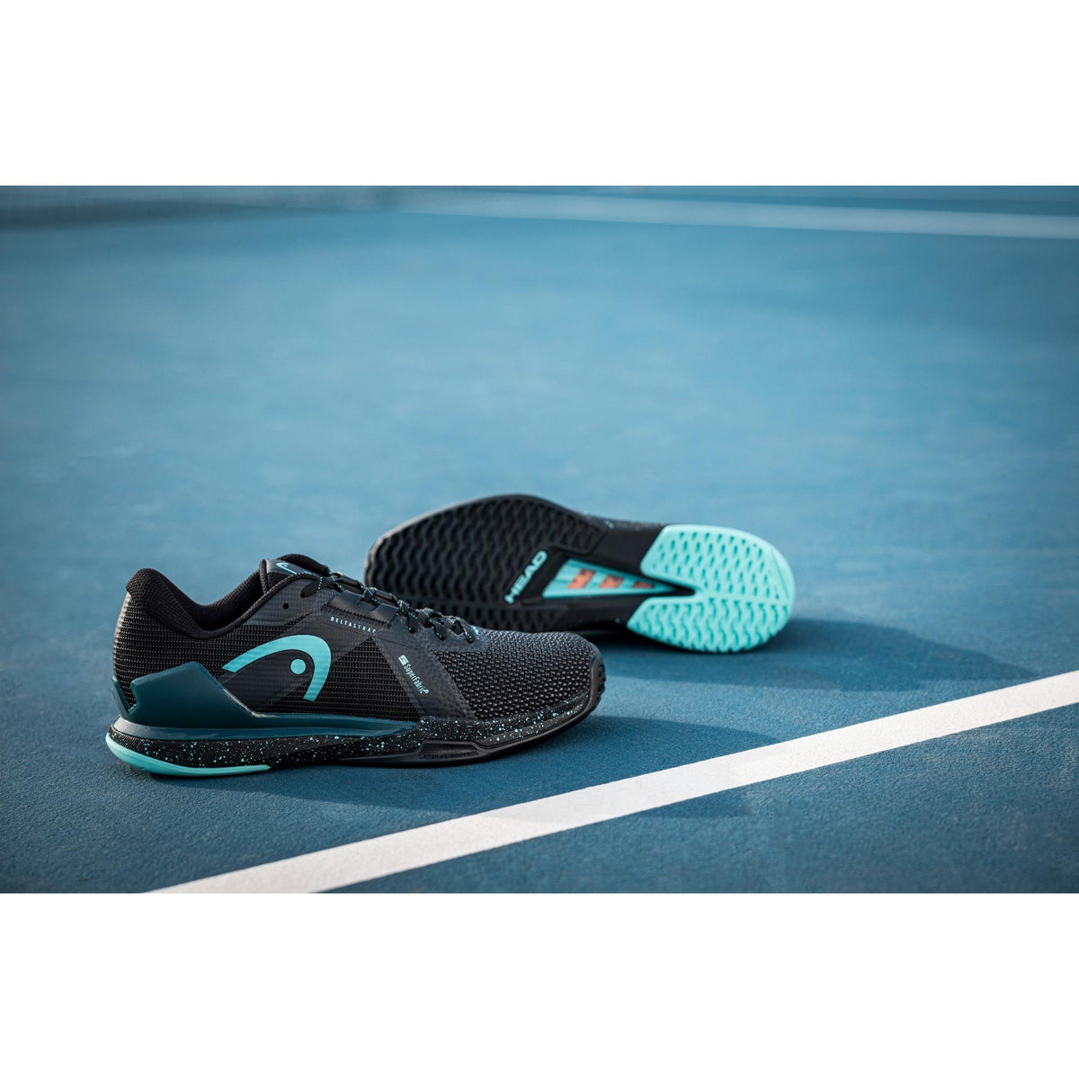 A pair of HEAD Sprint Pro 4.0 SF tennis shoes resting on a blue hard court, with one shoe upright and the other flipped to show the teal-accented outsole, emphasizing the lightweight mesh upper, low-profile design, and traction built for fast, responsive court movement.