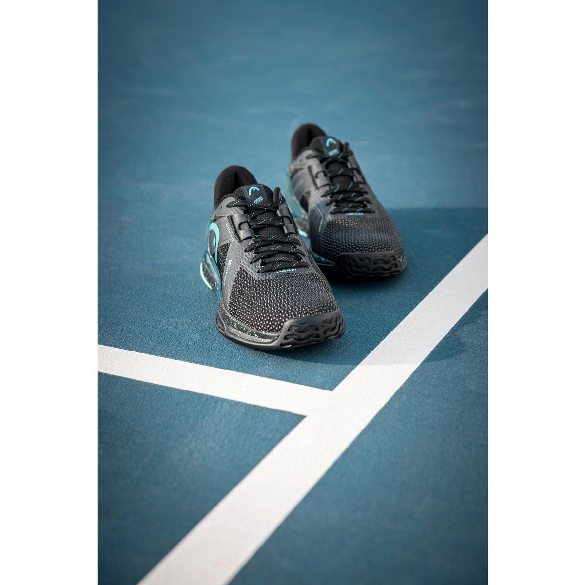 A pair of HEAD Sprint Pro 4.0 SF tennis shoes positioned at the intersection of white court lines on a blue hard court, shot from a low front angle to highlight the breathable knit upper, streamlined silhouette, and teal performance accents designed for speed, agility, and precision footwork.