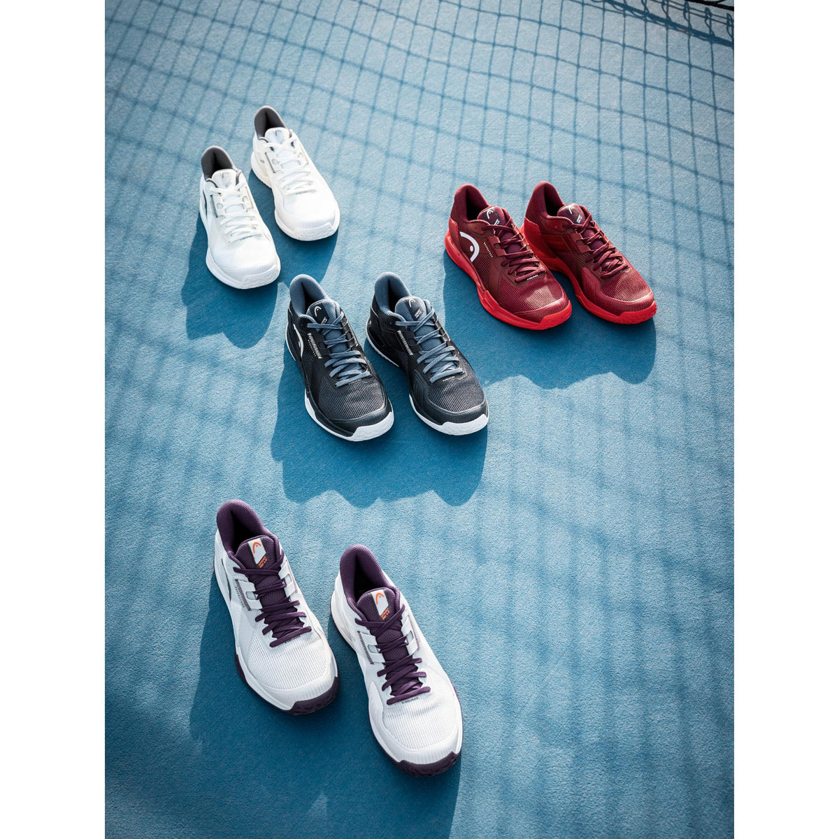 A top-down lifestyle image of multiple pairs of HEAD Sprint Pro tennis shoes arranged on a blue hard court. The shoes are displayed in several colorways—white, black, red, and white with purple accents—spaced evenly across the court surface. Soft natural sunlight casts long shadows, while the faint pattern of a tennis net falls across the background, highlighting the variety of designs, clean silhouettes, and performance-focused styling of the Sprint Pro c