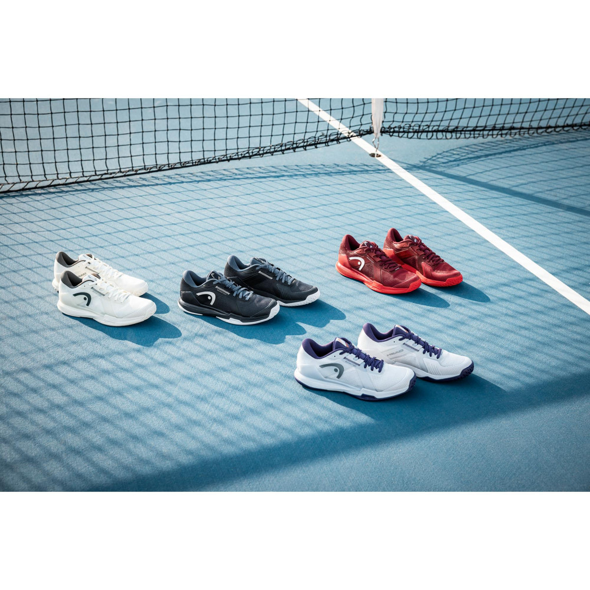 A wide lifestyle shot of multiple pairs of HEAD Sprint Pro tennis shoes neatly arranged on a blue hard court near the net. Five colorways are displayed—white, black, red, and white with purple accents—spaced evenly in a clean horizontal layout. Bright natural sunlight casts crisp shadows, while the tennis net creates a subtle grid pattern across the court surface. The composition highlights the range of color options, court-ready performance design, and sleek, modern styling of the Sprint Pro lineup in a ca