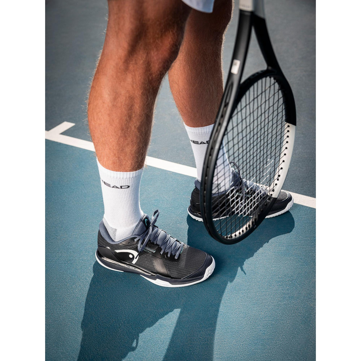 A close-up lifestyle shot of a tennis player standing on a hard court, wearing HEAD Sprint Pro tennis shoes. The image focuses on the lower legs and feet, highlighting the shoe’s streamlined black and grey knit upper, supportive side branding, and white non-marking outsole. A HEAD tennis racket rests near the player’s leg, reinforcing the on-court performance context. The lighting and shadows emphasize stability, grip, and a lightweight, speed-focused design built for competitive play.
