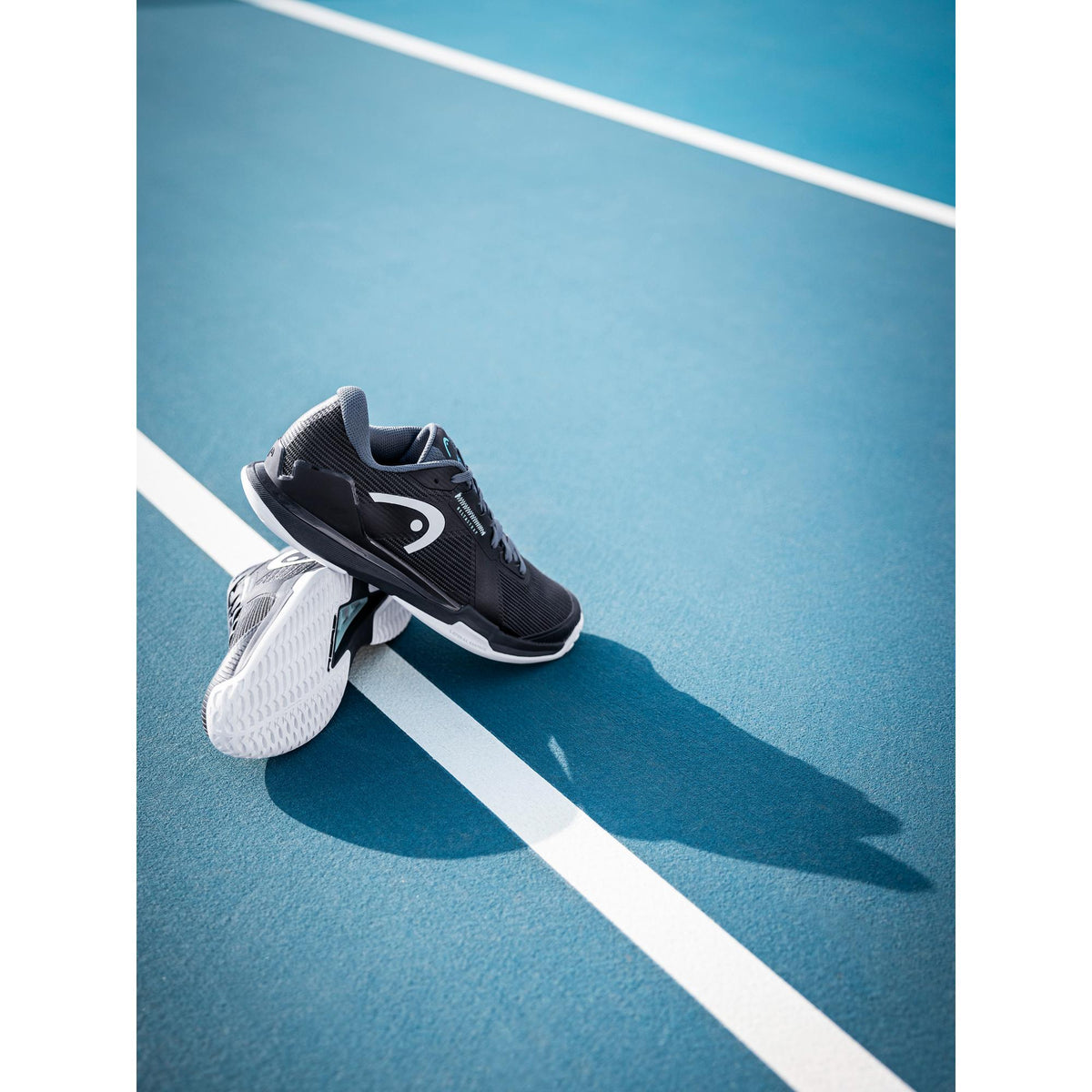 A clean lifestyle product shot of the HEAD Sprint Pro tennis shoes resting on a hard court. One shoe is upright while the other is angled to showcase the white non-marking outsole, with crisp court lines cutting diagonally through the frame. The black and grey upper, streamlined silhouette, and bold side branding stand out against the blue surface, while natural sunlight casts sharp shadows that emphasize grip, stability, and a lightweight, speed-driven design built for fast, aggressive movement on court.