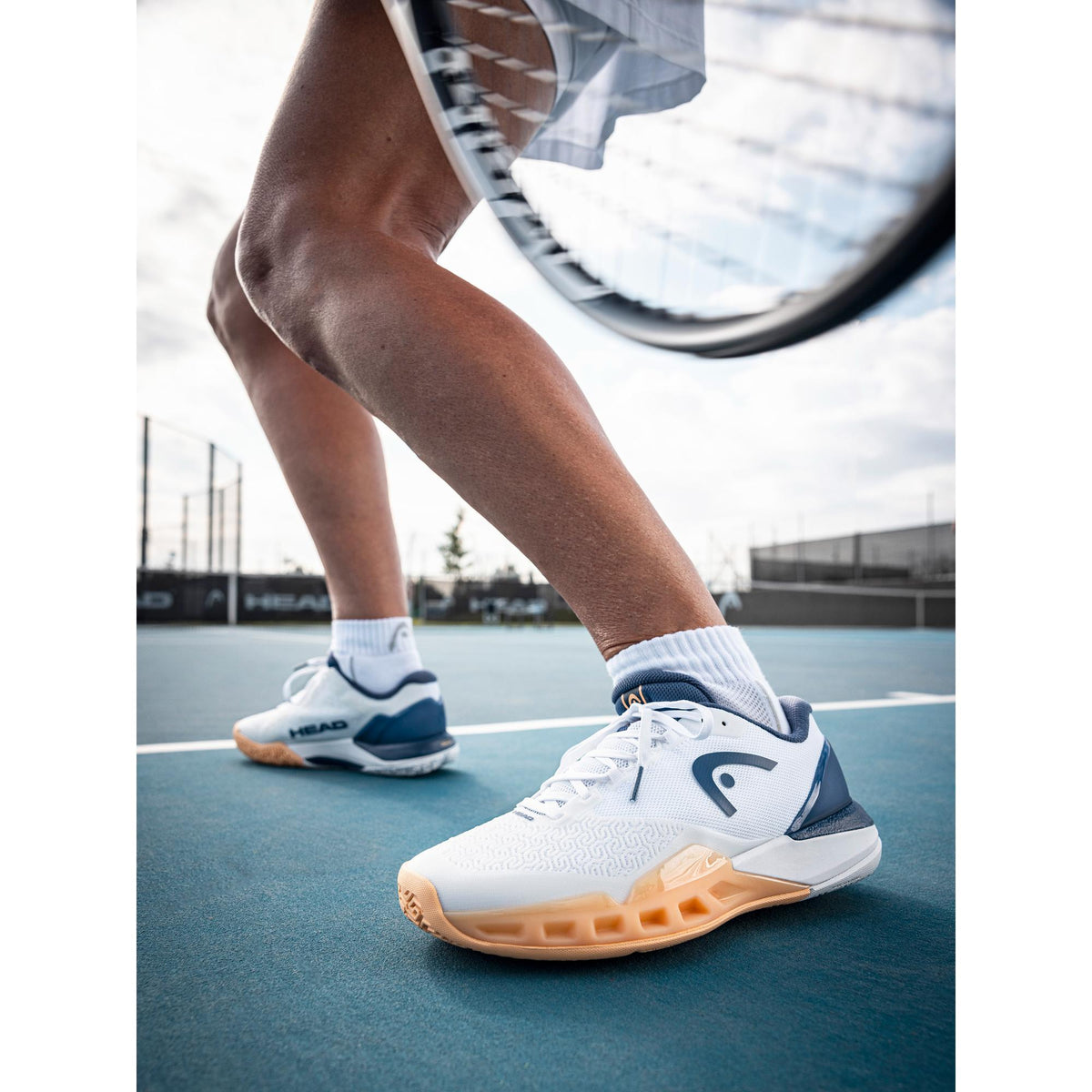 Close-up lifestyle shot of a tennis player stepping forward on a blue hard court while wearing white HEAD Revolt Pro tennis shoes with navy accents and a gum outsole, captured mid-movement with a racket in motion in the background.