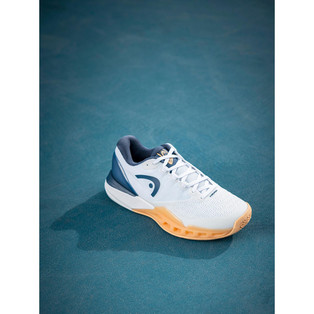 Product-focused lifestyle image of a single white HEAD Revolt Pro tennis shoe with navy detailing and a gum outsole placed on a blue hard court, shown from a three-quarter angle with soft natural lighting and a clean, minimal background.
