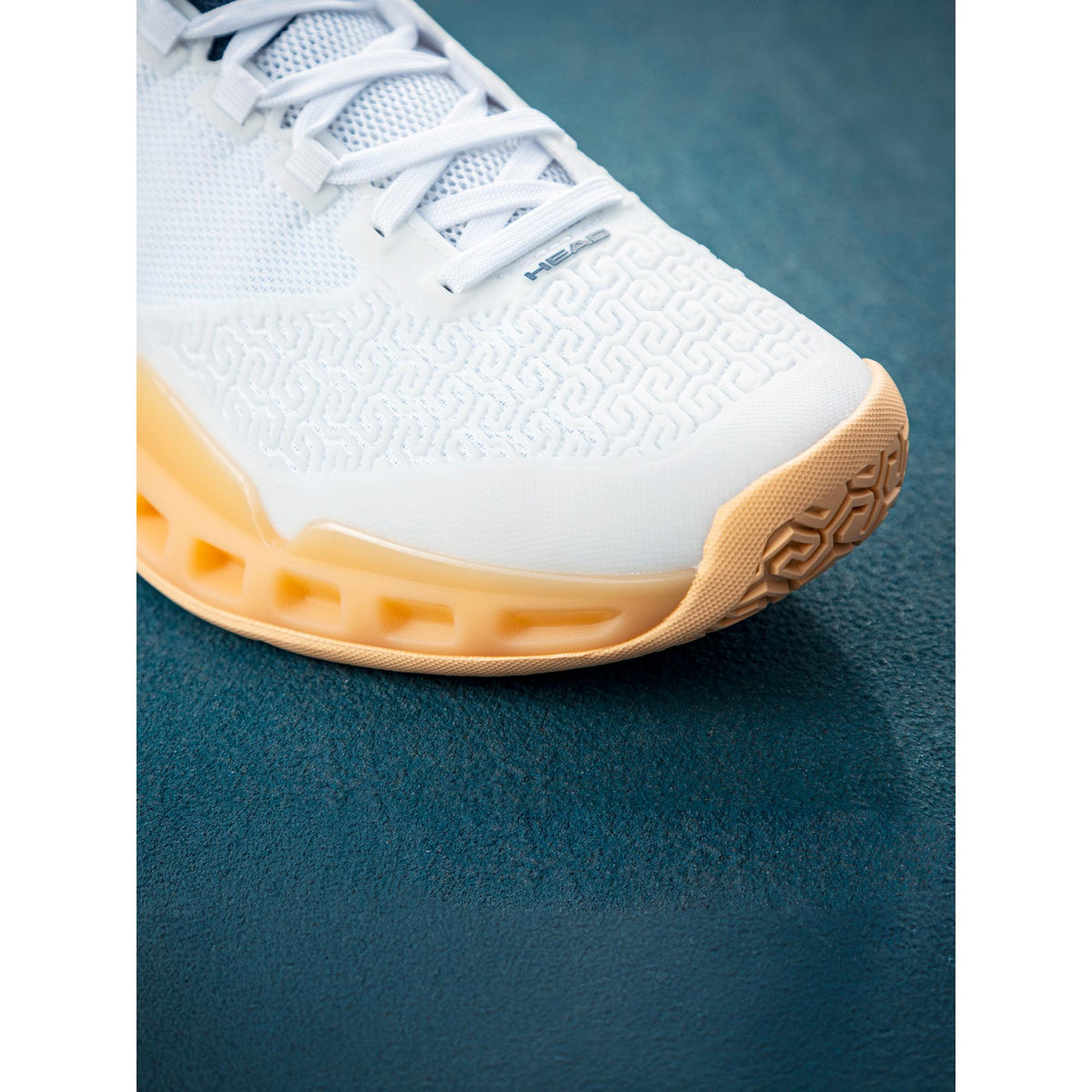 Close-up detail shot of the toe and forefoot of a white HEAD Revolt Pro tennis shoe, highlighting the textured upper, white laces, and gum rubber outsole on a blue hard court surface under natural light.