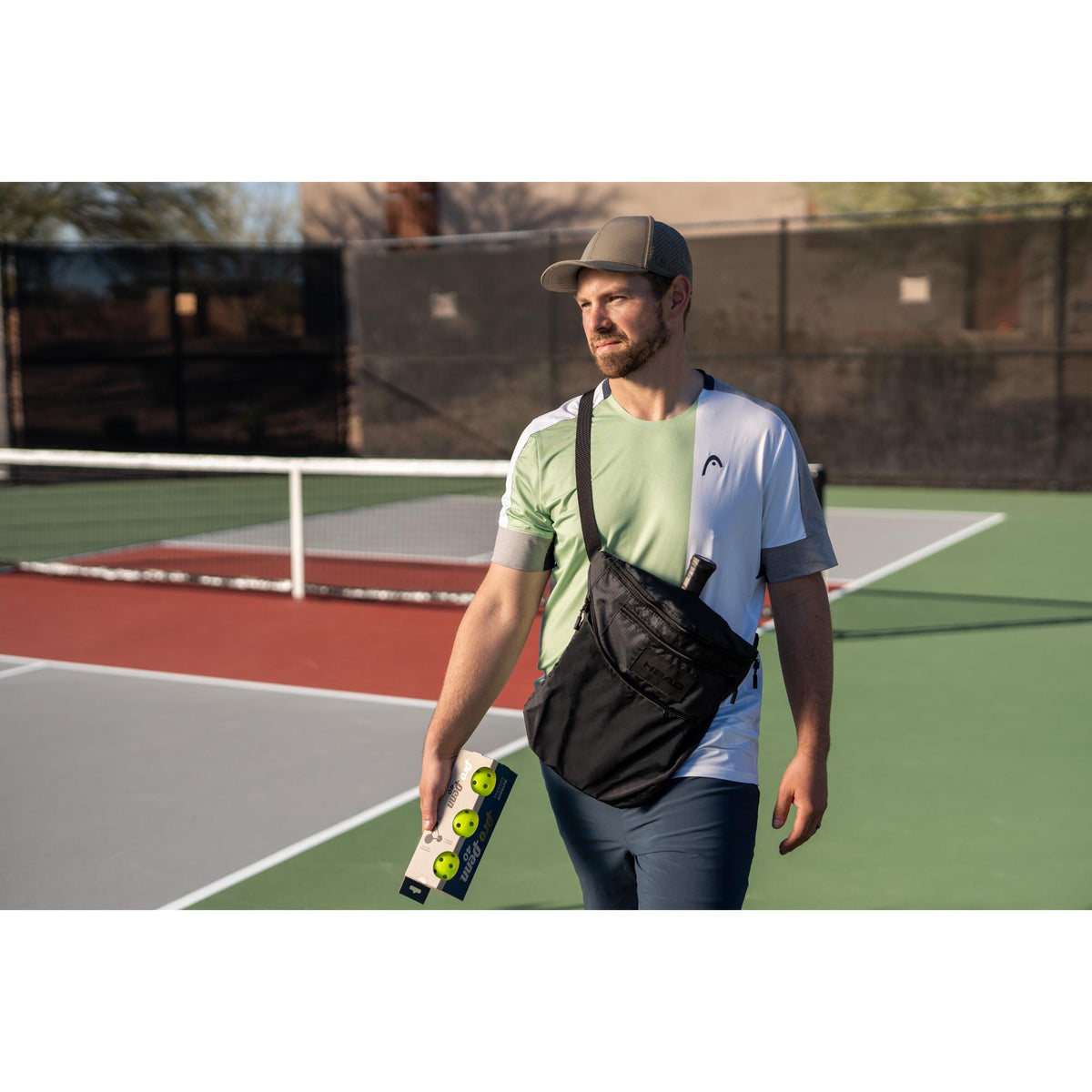Man wearing a black HEAD tour crossbody bag walks across an outdoor pickleball court holding a pack of pickleballs, with a paddle handle visible in the bag and a net and courts in the background.