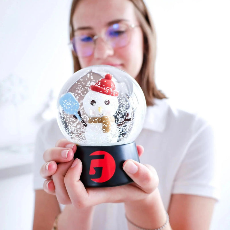 Snow globe featuring a snowman holding a paddle inside a clear dome with falling snow effect, mounted on a display base.