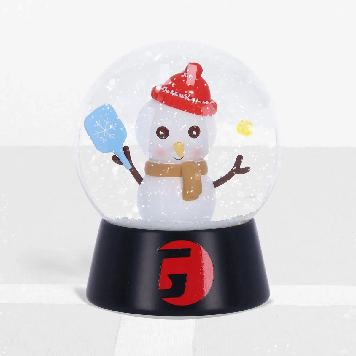 Snow globe featuring a snowman holding a paddle inside a clear dome with falling snow effect, mounted on a display base.