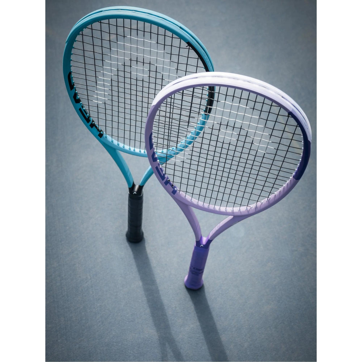 Two HEAD Boom TEAM tennis racquets in teal and lavender standing crossed on an outdoor tennis court.