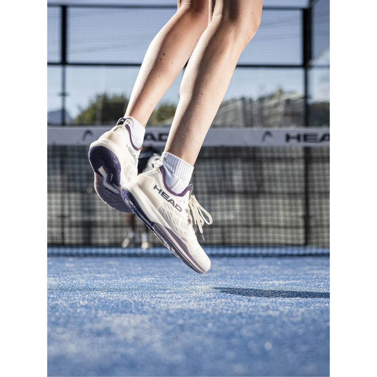 Female padel player mid-jump wearing HEAD Motion Pro 1.5 women’s padel shoes in off-white with plum accents on a blue padel court, captured from a low angle to highlight the outsole grip, heel cushioning, and HEAD branding during dynamic movement.