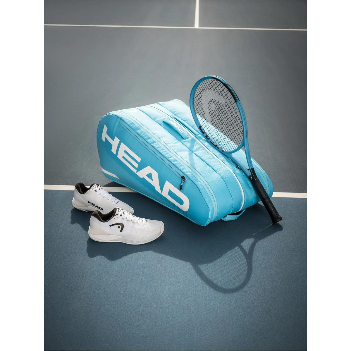 HEAD Boom TEAM tennis racquet leaning against a teal HEAD tennis bag with white shoes on a tennis court.
