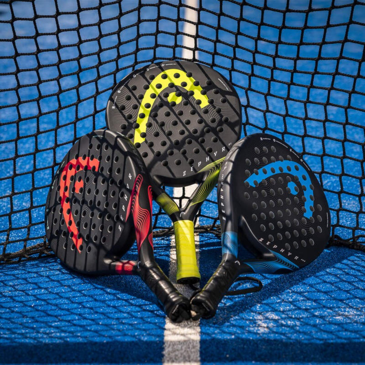 Three HEAD Zephyr padel rackets in red, yellow, and blue colorways arranged on a blue court in front of a net.