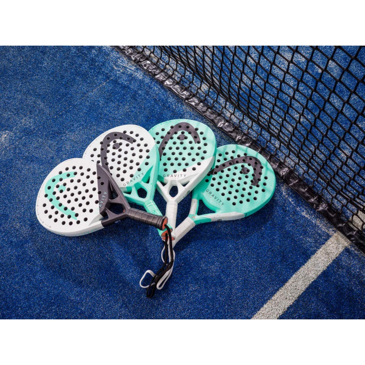 Four HEAD Gravity padel rackets on blue court beside net, displayed in white and mint colorways.