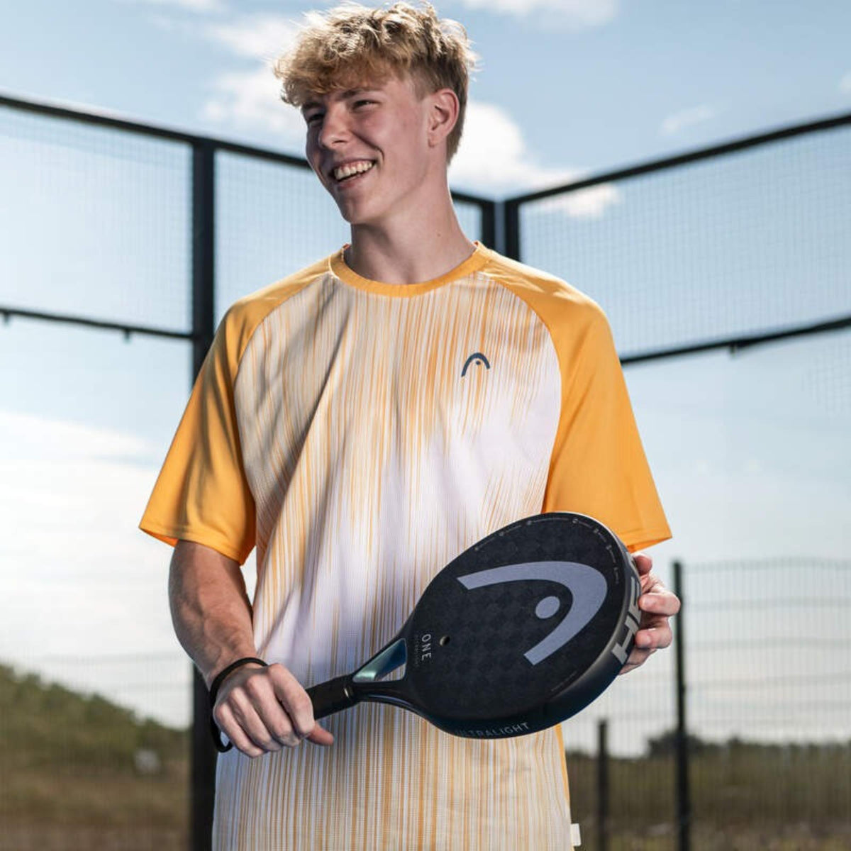 Young male player holding Head One Ultralight padel racket on an outdoor court, smiling.