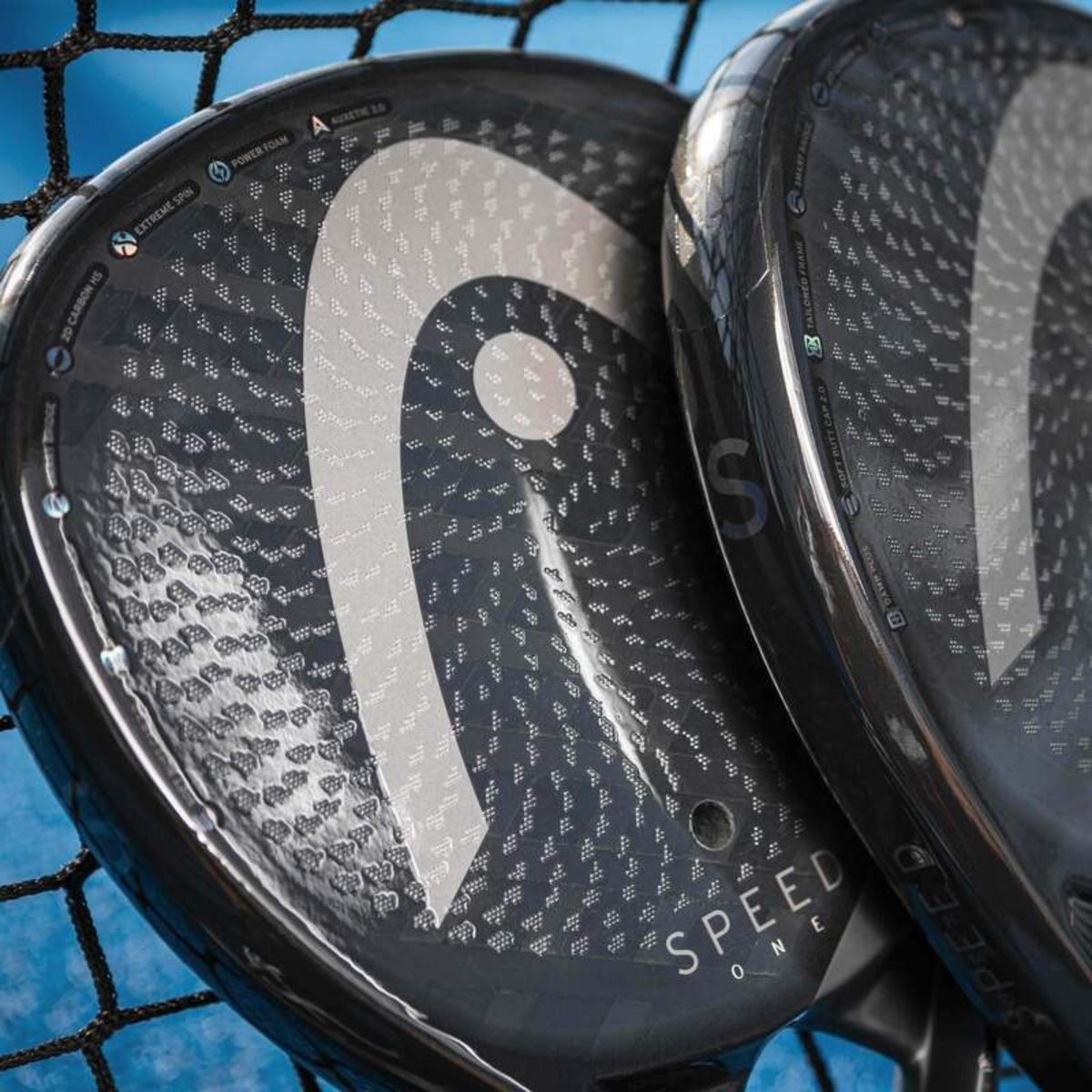 Two HEAD Speed One padel rackets resting against padel net showing textured carbon surface and logo.