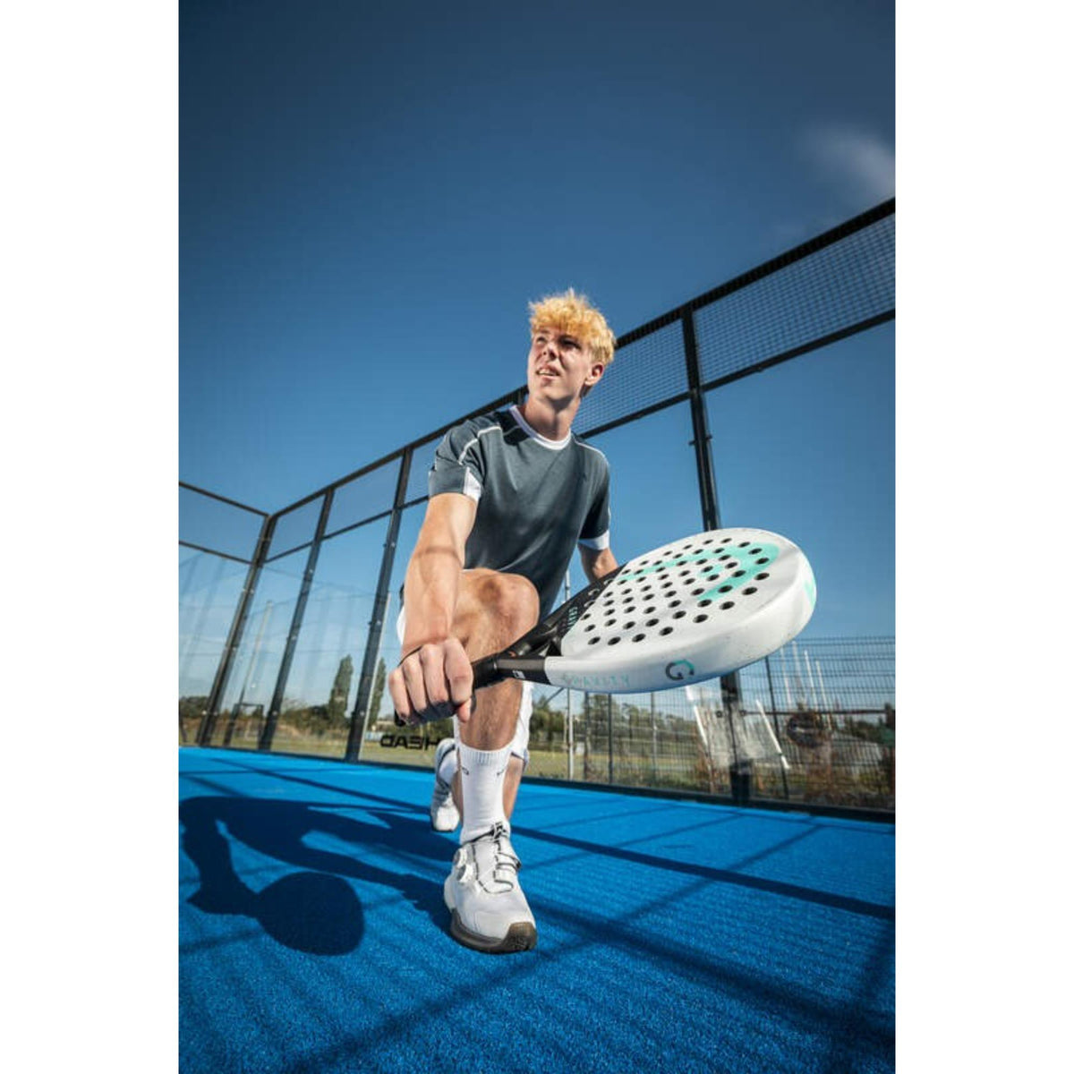 Young male athlete playing padel with HEAD Gravity Pro racket on outdoor blue court under clear sky.