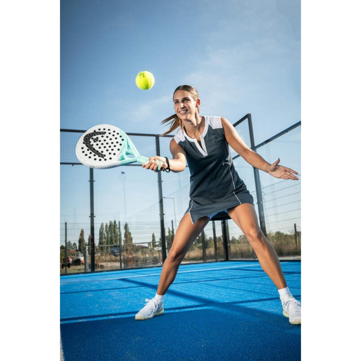 Female player hitting ball with Head Gravity padel racket on outdoor blue padel court during play.